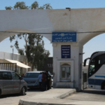 Border checkpoint entrance with a blue 'Arrivals and Departures' sign, a VIP desk, and a large bus on the right amid parked cars.