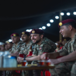 Group of soldiers in camouflage uniforms and maroon berets seated at a long table at a dimly lit event, smiling and watching something off-camera.