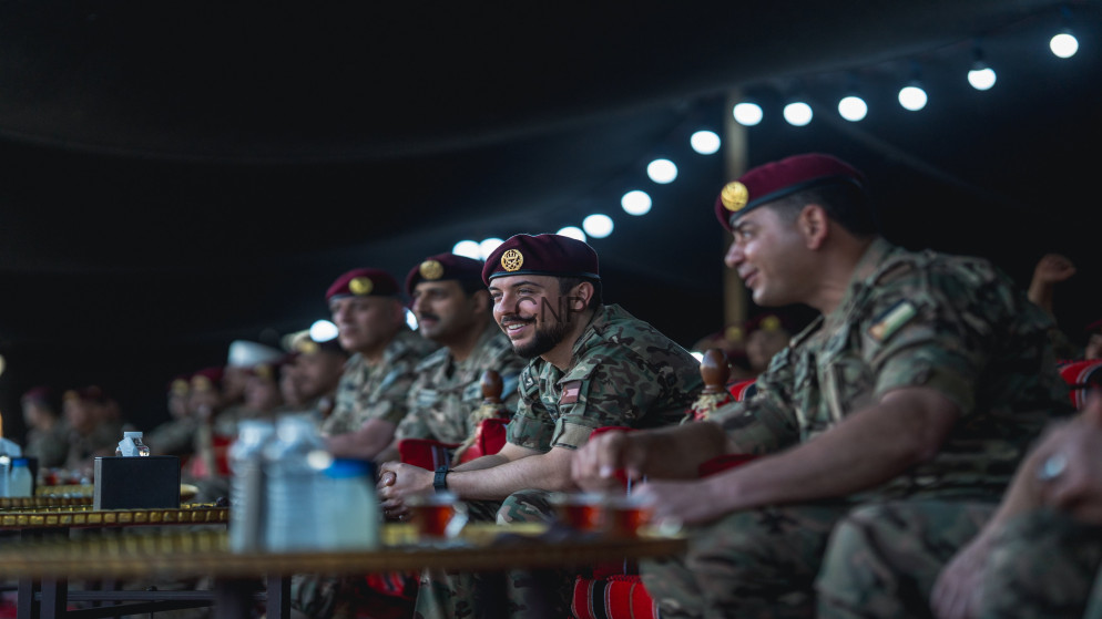 image - Center News Network Group of soldiers in camouflage uniforms and maroon berets seated at a long table at a dimly lit event, smiling and watching something off-camera.