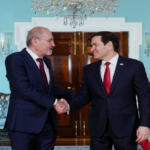 Two suited men shake hands in a formal room, flanked by the Jordanian and American flags behind them.