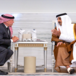 Two men sit in ornate chairs across a small table, wearing traditional Middle Eastern attire, conversing in a marble-floored room.