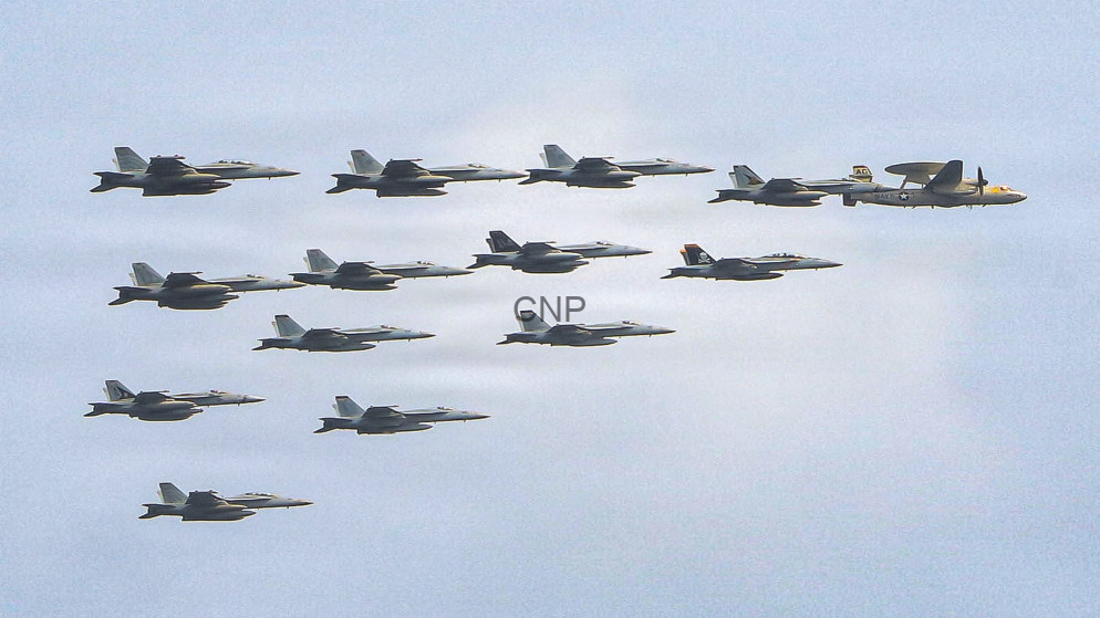 Twelve fighter jets flying in a tight formation across a clear blue sky, with a larger support aircraft at the far right.