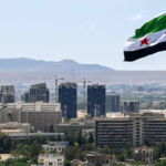 City skyline with a large Syrian flag on a tall pole in the foreground and construction cranes behind, under a clear blue sky and distant hills.