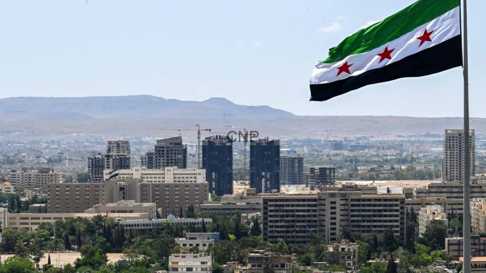 image - Center News Network City skyline with a large Syrian flag on a tall pole in the foreground and construction cranes behind, under a clear blue sky and distant hills.