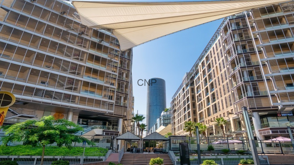 image - Center News Network Open-air plaza framed by two curved beige buildings, with a tall blue glass tower in the background and shaded seating areas amid palm trees and greenery.