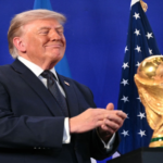 Donald Trump in a suit, smiling and clapping at a podium, with American flags in the background and a gold trophy nearby.
