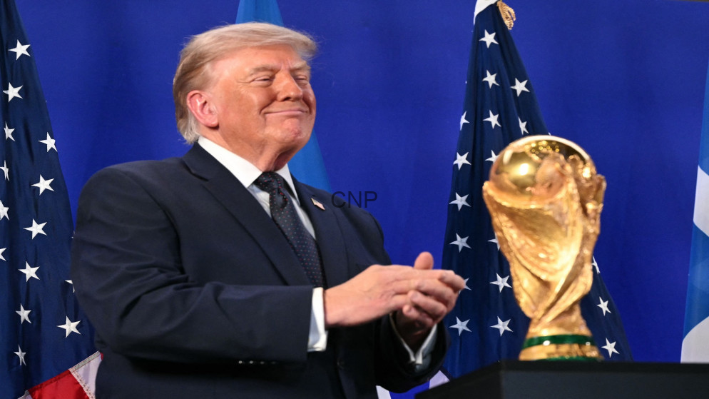 Donald Trump in a suit, smiling and clapping at a podium, with American flags in the background and a gold trophy nearby.