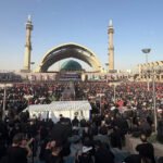 Large crowd gathered at a mosque plaza with a green-domed shrine and tall minarets during a religious event.
