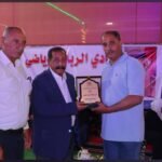 Three men on a stage during an award ceremony; one man in a suit presents a plaque to another in a light blue polo, with an Arabic banner behind them.