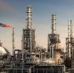 Oil refinery complex with tall smokestacks, distillation towers, and a network of pipes against a clear sky