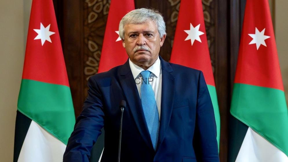 Man with gray hair and mustache in a navy suit and light blue tie stands at a podium, with Jordanian flags in the background.