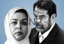 Two adults pose seriously against a blue backdrop; woman wearing a white headscarf and a man with dark hair and stubble.