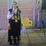 Woman in a black gown with gold embroidery holding a microphone at an indoor event, near a yellow chair and speaker rigging.