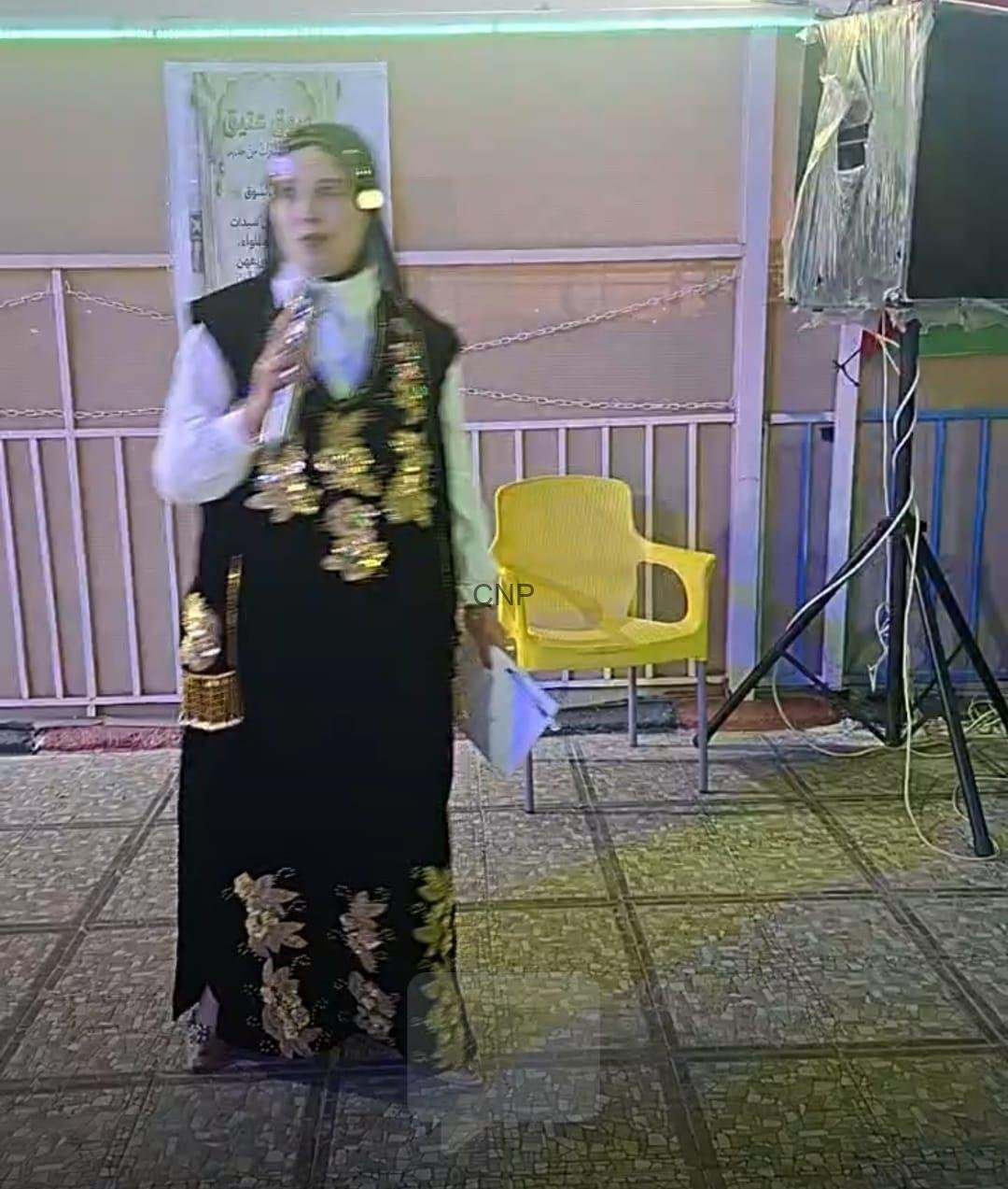 to2 - Center News Network Woman in a black gown with gold embroidery holding a microphone at an indoor event, near a yellow chair and speaker rigging.