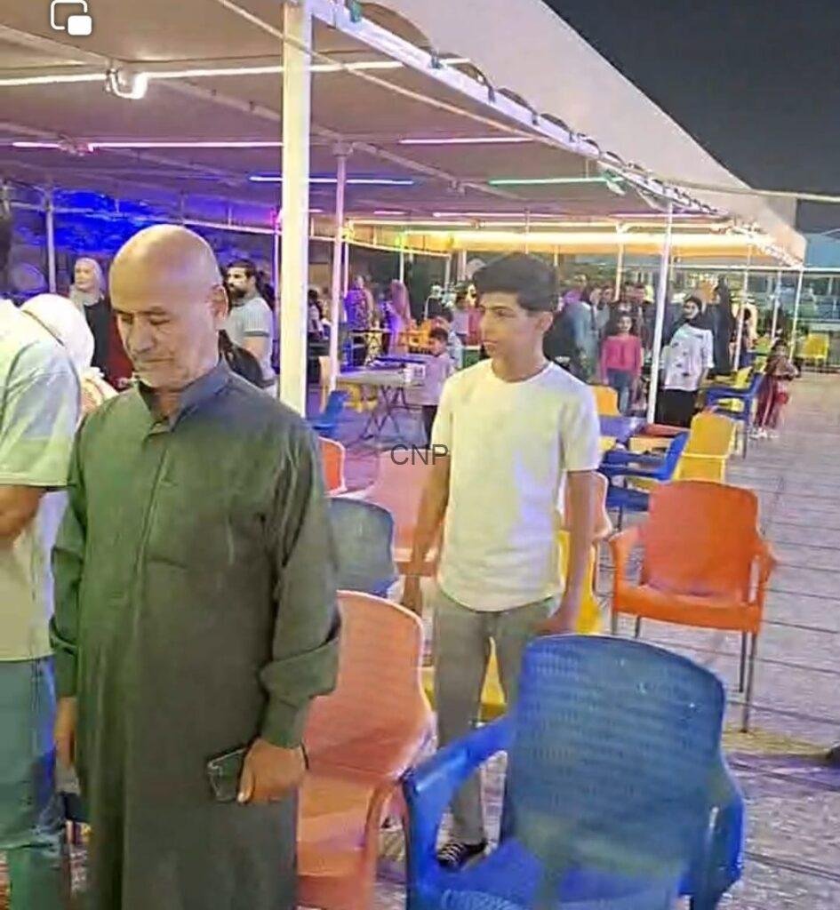 to5 - Center News Network Bald man in a green traditional kurta stands beside colorful plastic chairs at a nighttime outdoor event, holding a phone.