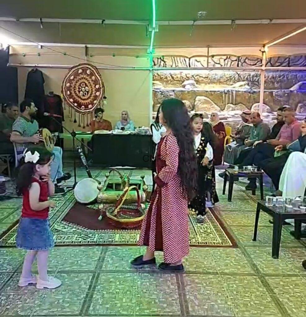 to6 - Center News Network Girl in a red patterned dress dances on a decorative rug while a live band and seated guests watch in a cultural gathering tent.