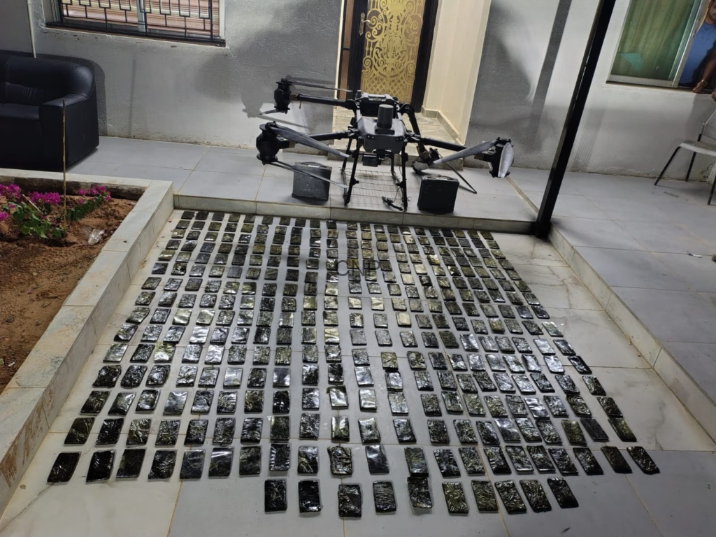 Drone rests on tiled patio beside a doorway, with hundreds of small rectangular black packages laid out in neat rows on the floor for inspection or inventory.