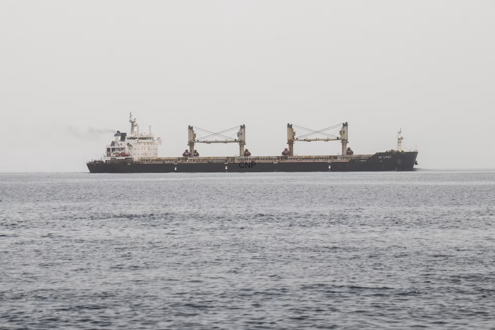 image - Center News Network Large cargo ship with two gantry cranes sailing on a calm, gray sea under a pale sky.