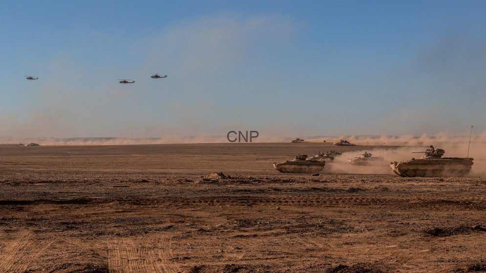 image - Center News Network Armored tanks advance across a dusty desert battlefield with helicopters flying overhead.