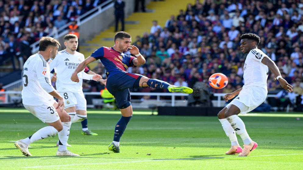 image - Center News Network Barcelona player in blue and maroon kicking the ball while Real Madrid players in white close in during a crowded match on a sunny day.
