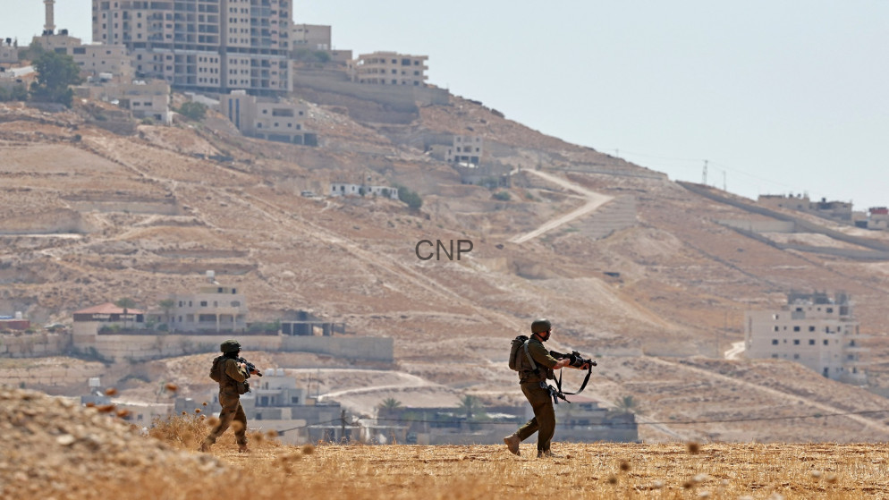 image - Center News Network Two soldiers in brown uniforms and helmets patrol a dry hillside with rifles as a town climbs the hill in the background.