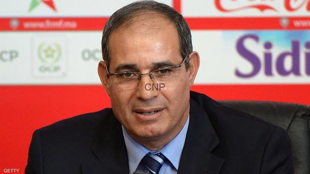 Close-up of a middle-aged man in a dark suit and blue striped tie, wearing glasses, speaking at a press conference with a red backdrop and logos behind him.