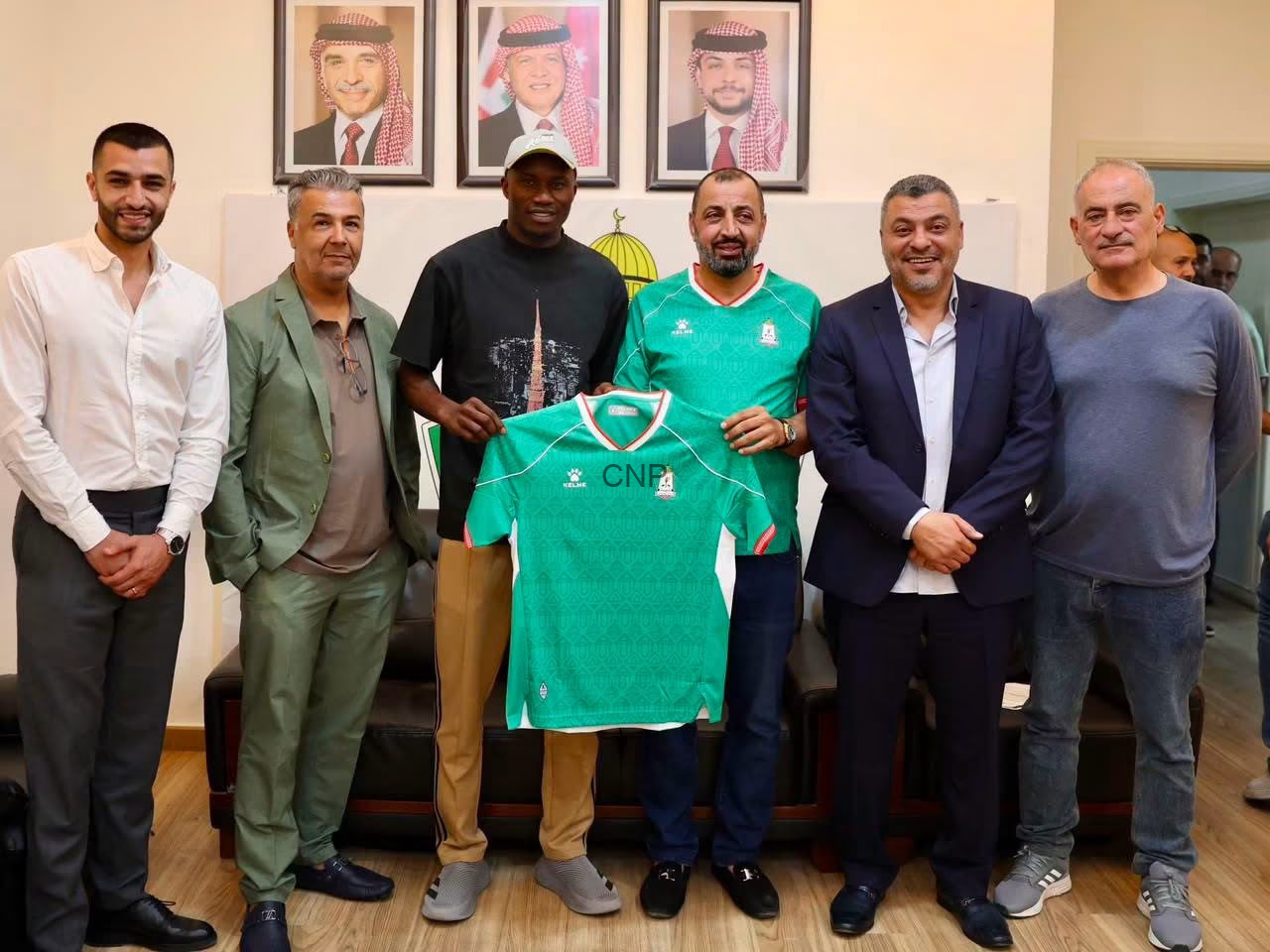 Seven men standing indoors, smiling, while one holds a green sports jersey in front of them.