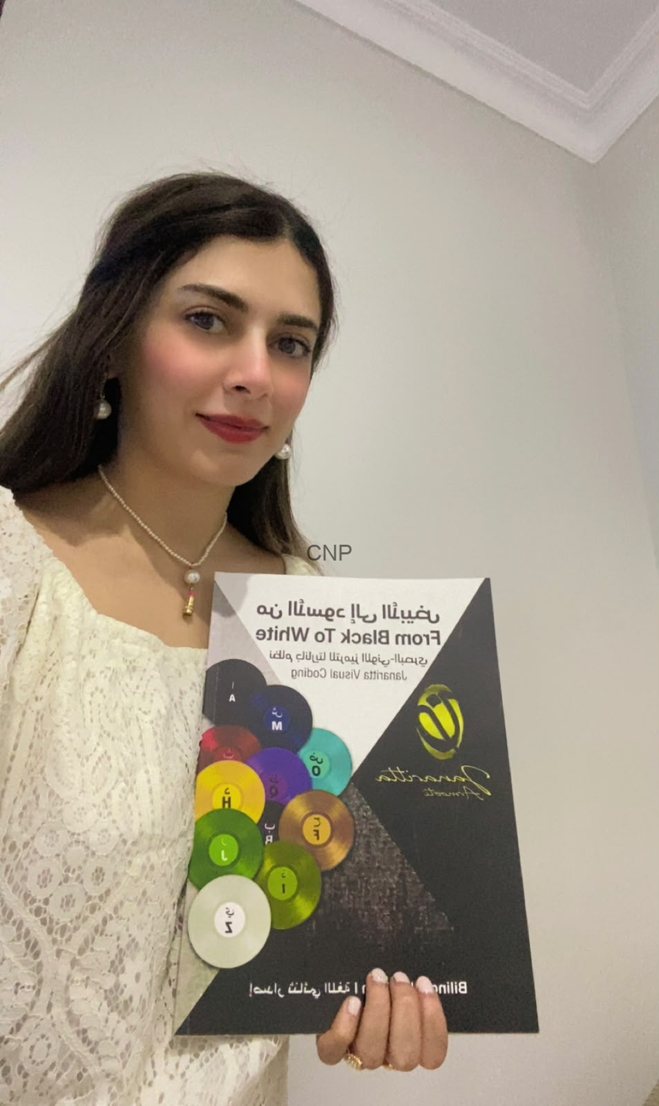 Young woman wearing a white lace top and pearl necklace holds a colorful brochure with circular color swatches and Arabic text on the cover.