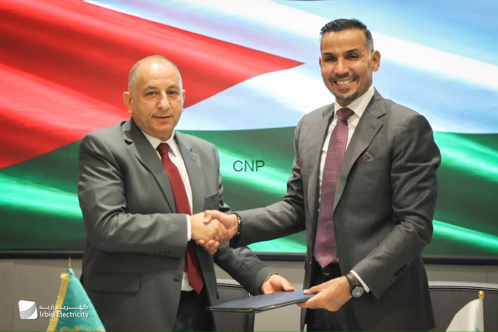 image - Center News Network Two men in suits shake hands over a signed document, with a Jordanian flag backdrop and Irbid Electricity logo nearby, symbolizing a formal agreement.