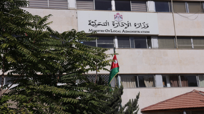 Front view of a government building with a sign reading Ministry of Local Administration and a Jordan flag in front, partially shaded by trees.