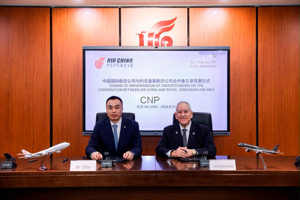 image - Center News Network Two executives in suits sign a memorandum at a polished wooden conference table, with model airplanes and a screen showing Air China and Royal Jordanian Airlines logos in the background.