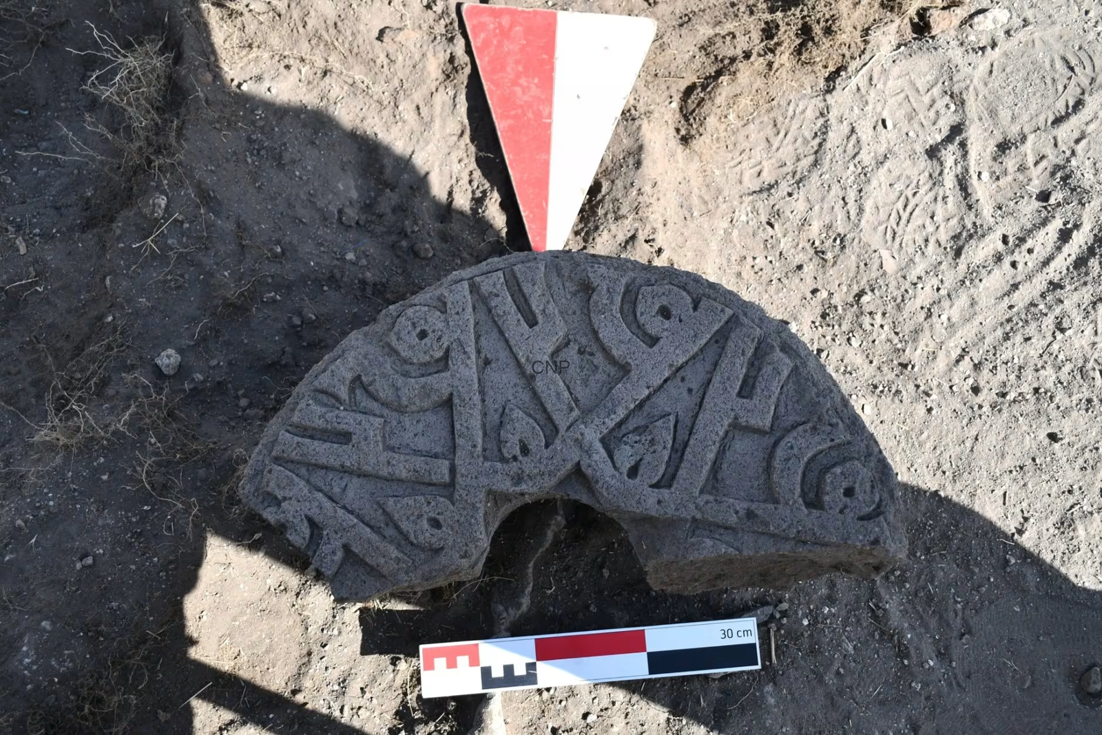 Circular, carved stone manhole cover half-buried in dirt, with a red-and-white survey marker and a 30 cm scale bar nearby.