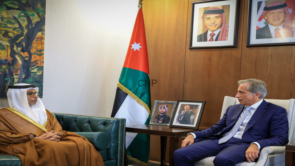 image - Center News Network Two men sit in a formal office: a traditional-dressed Jordanian elder on a green leather sofa and a suited man on a chair, with a Jordan flag and portraits behind them.