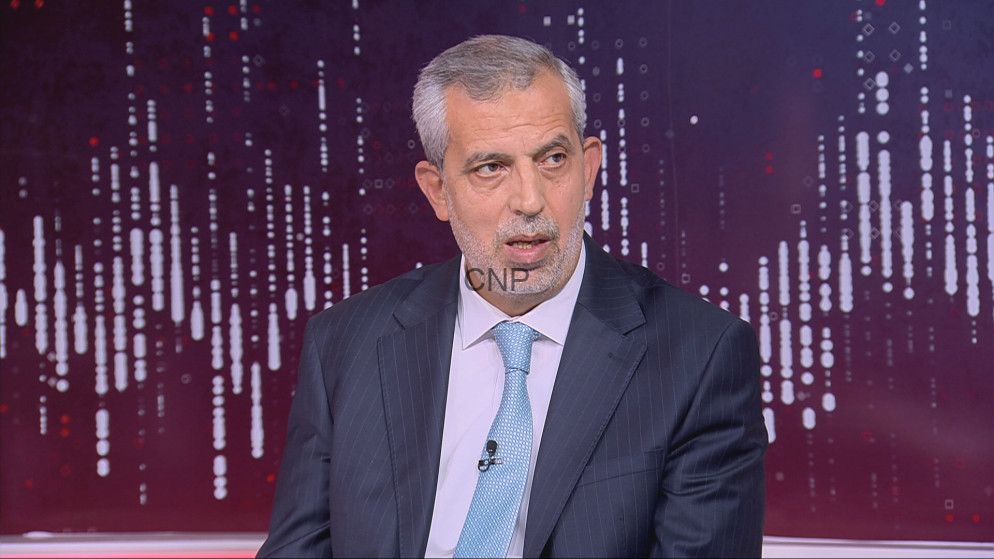 image - Center News Network Man in a dark pinstripe suit and light blue tie seated in a TV studio during an interview, microphone clipped to the tie, purple abstract background.
