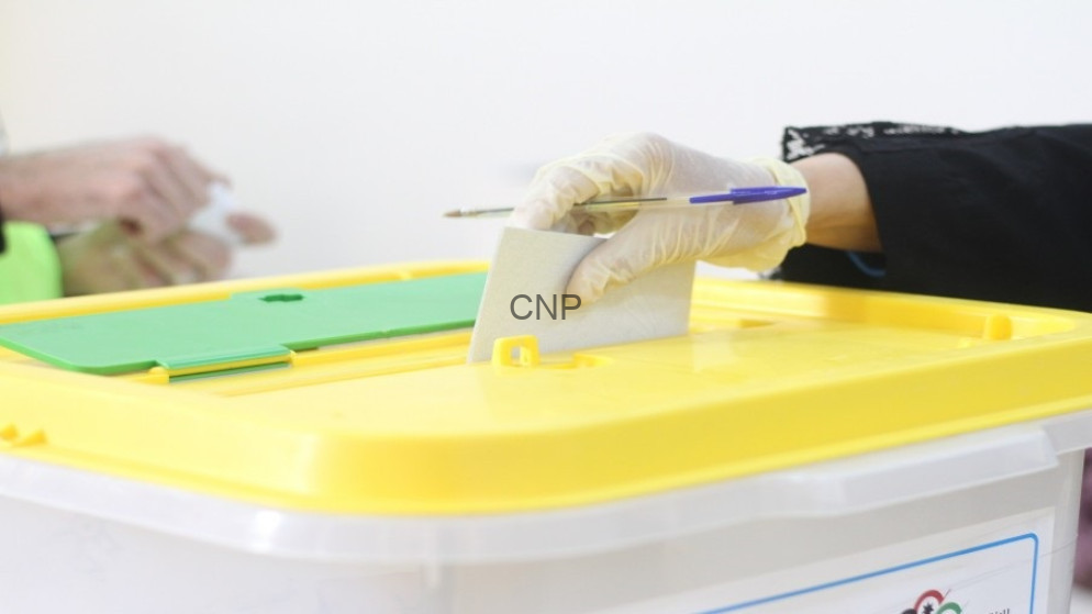 image - Center News Network Person wearing gloves inserts a ballot into a yellow ballot box during voting.