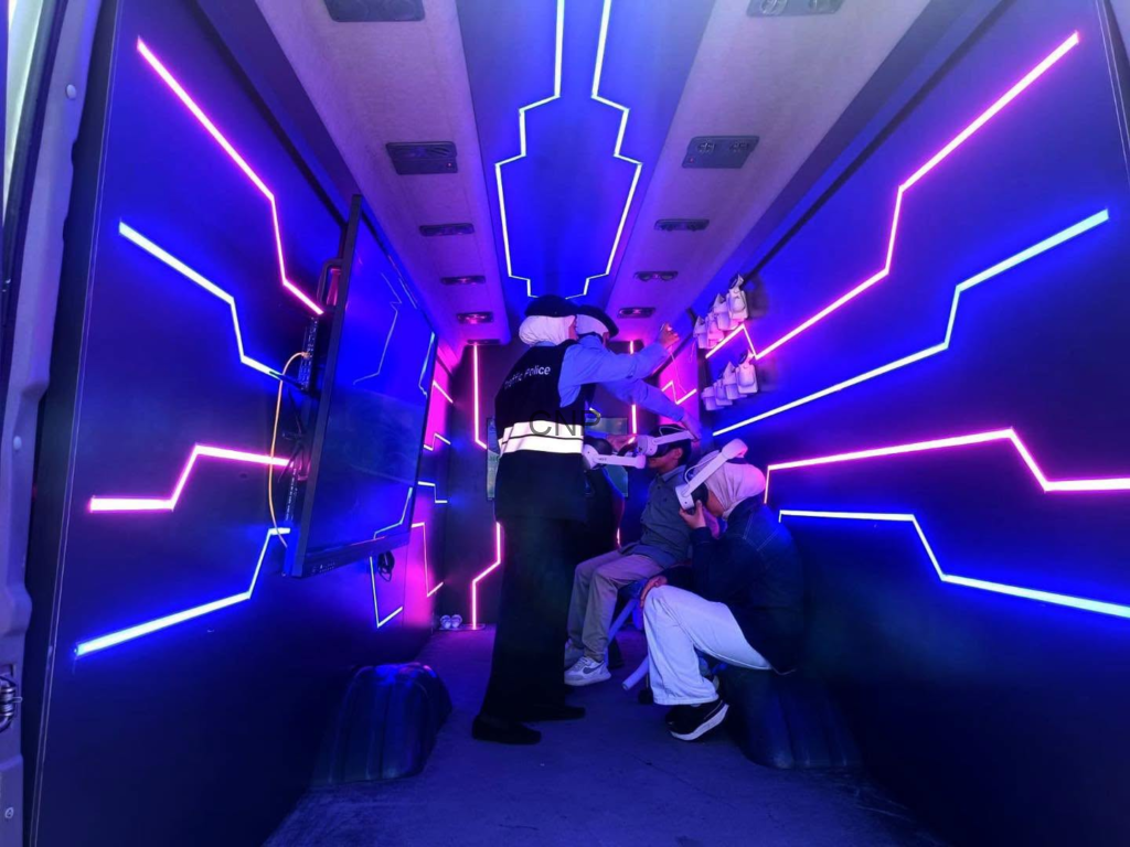 image - Center News Network Inside a neon-lit vehicle, a uniformed officer in a reflective vest talks to two seated passengers while others stand nearby.
