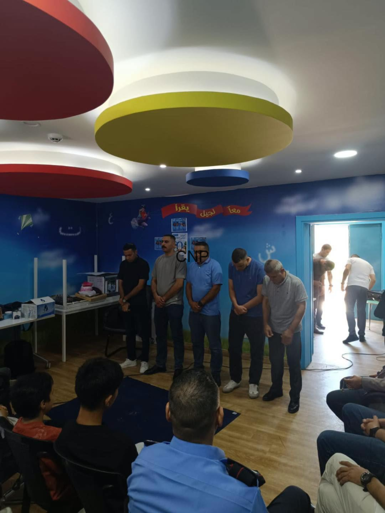 image - Center News Network Five men stand with heads bowed in a line inside a bright blue room, audience seated and observing, colorful circular ceiling lights overhead.