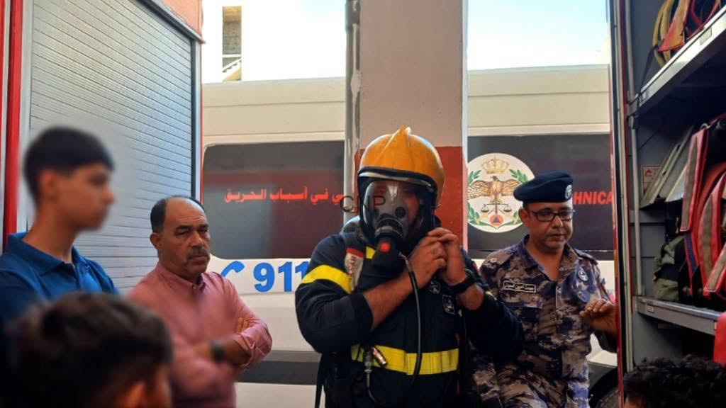 Firefighter in a yellow helmet and gas mask speaks into a microphone beside an ambulance, while a uniformed officer and bystanders look on.