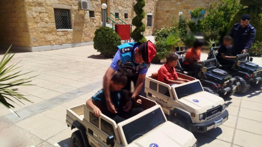 image - Center News Network Police officer in uniform helps children ride in small plastic toy SUVs in a sunny courtyard with beige stone buildings and greenery.