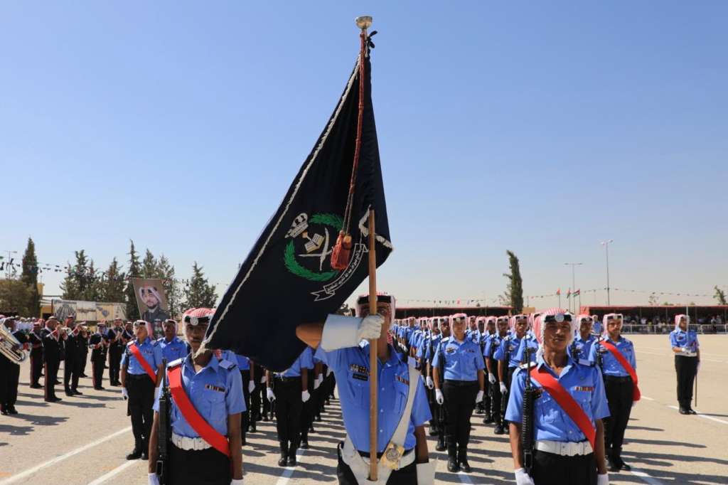 Color guard leading a parade: flag bearer holding a large black banner with emblem, surrounded by rows of uniformed troops in blue shirts and red sashes on a sunny day.