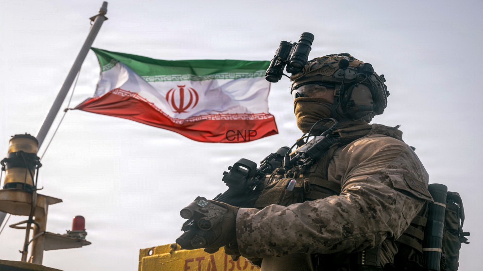 image - Rakeen News Armed soldier in desert camouflage with night-vision goggles, standing beside an Iranian flag.
