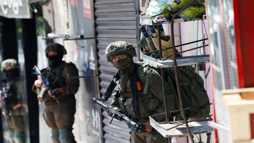 image - Center News Network Armed soldiers in tactical gear on a city street, one peering from behind a metal cart stacked with bags adjacent to a shuttered storefront.