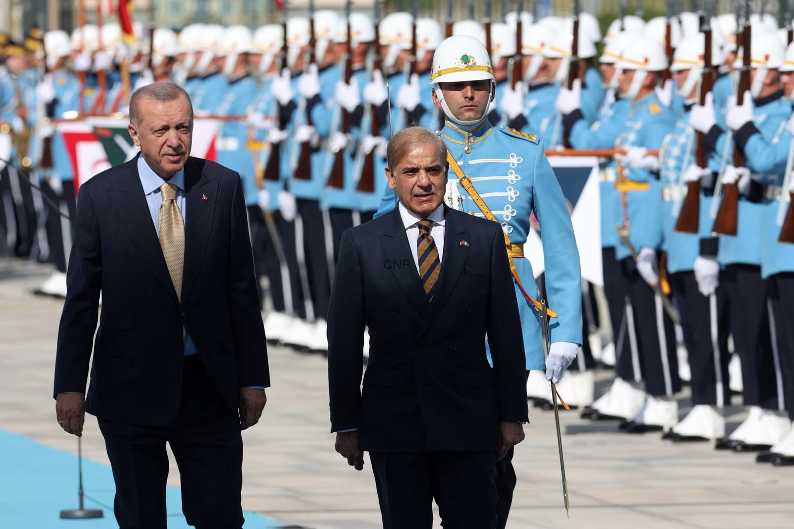 image - Center News Network Two men in dark suits walk in front of a ceremonial military parade, with soldiers in blue dress uniforms standing at attention in the background.