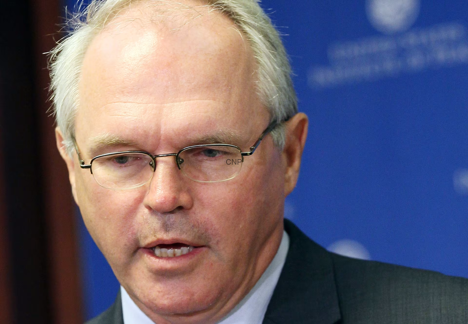 image - Center News Network Close-up of a man with gray hair and glasses in a dark suit, speaking at a press event against a blue backdrop.