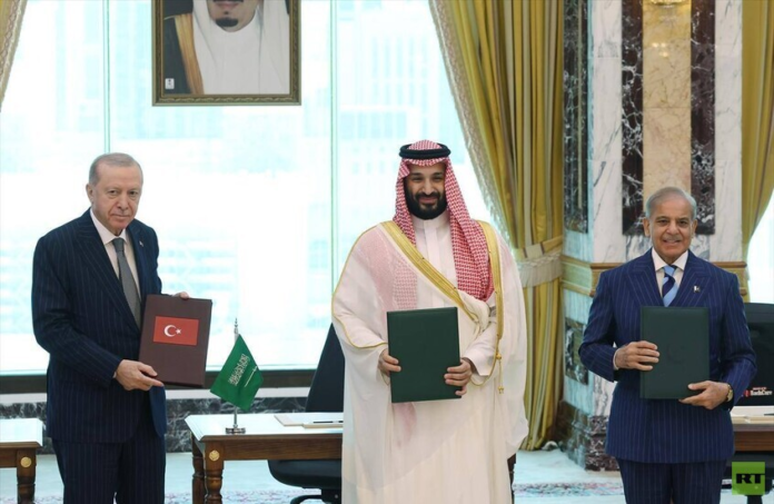 Three men in formal attire hold folders during a signing ceremony; Turkish and Saudi flags on the table, ornate room backdrop.