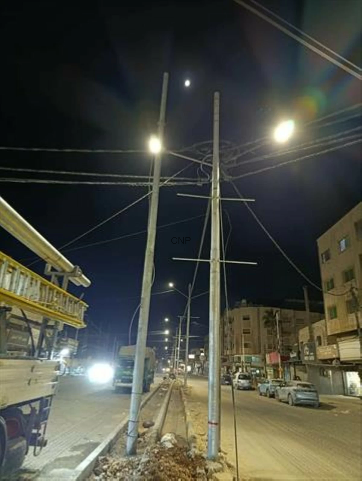 image - Center News Network Night street scene with tall utility poles and overhead power lines, glowing streetlights, and parked cars along the road.