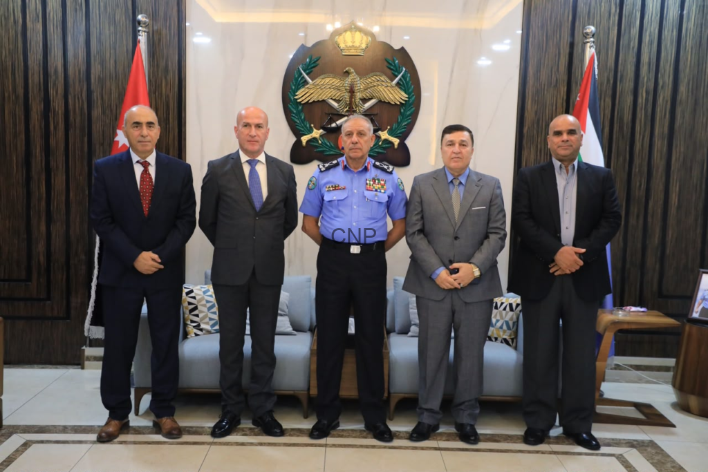 Five men in formal attire pose for a group photo in a government lobby, center officer in blue uniform with medals, emblem and flags in the background.