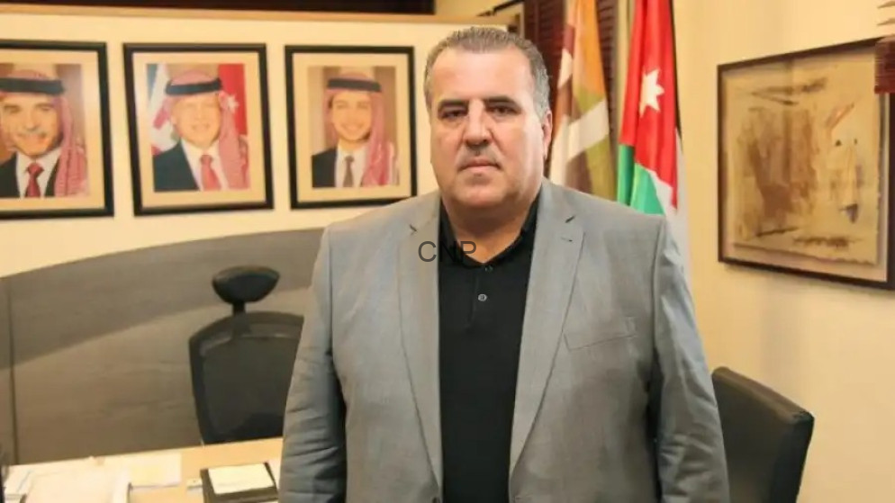 image - Center News Network Man in a gray suit stands in an office with framed portraits on the wall and flags in the background.