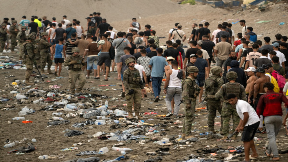 Armed soldiers stand on a litter-strewn beach as a crowd of civilians forms a long line behind them.