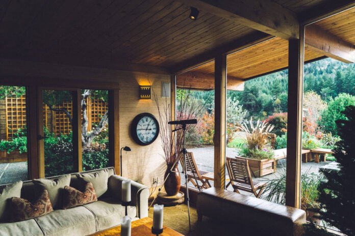 Sunlit living room with a beige sofa, candles on a coffee table, and large glass walls opening to a lush garden.